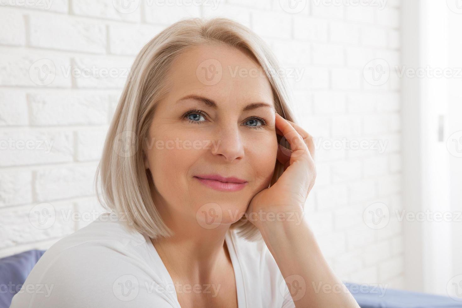 Positive woman with pillow sitting in bed and smiling to camera at home photo