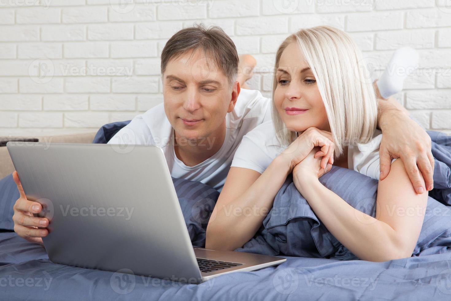 Smiling attractive couple lying on bed using laptop communicating online at home, happy man and woman typing on computer, enjoying internet shopping or chatting in social networks in bedroom. photo