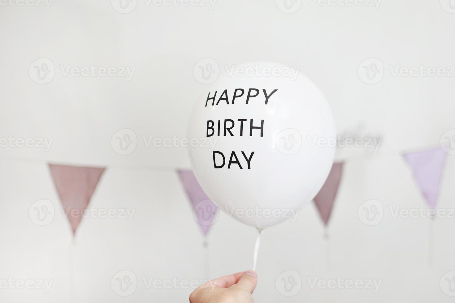 Unrecognised hand holding a white balloon with black inscription Happy birthday in decoration for the holiday. colourful garland flags at wall on background photo