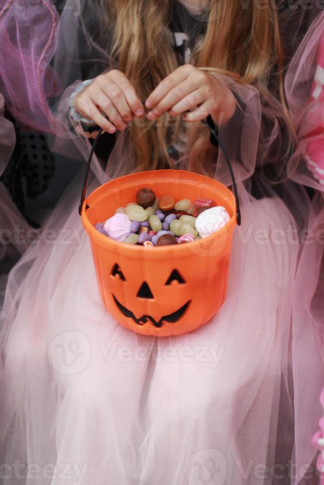 Cropped photo of child girl in a witch fancy dress holding a pumpkin-shaped candy basket with scary face in her hands. trick or treat. Different sweets. Concept of Halloween family holiday, festival