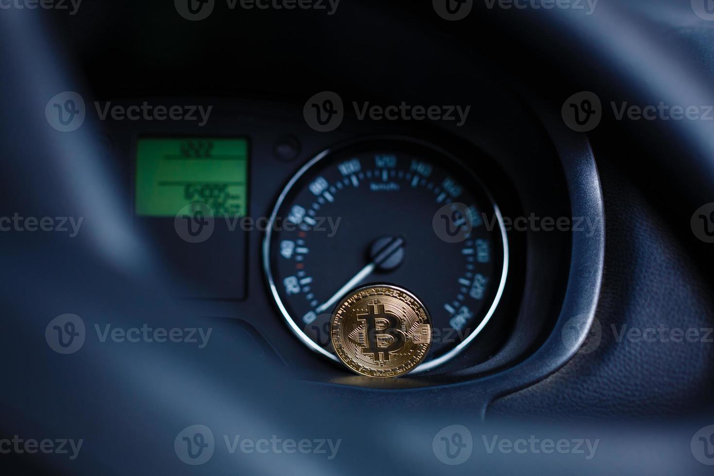 The physical coin is a gold bitcoin on the dashboard of the car next to the fuel consumption photo