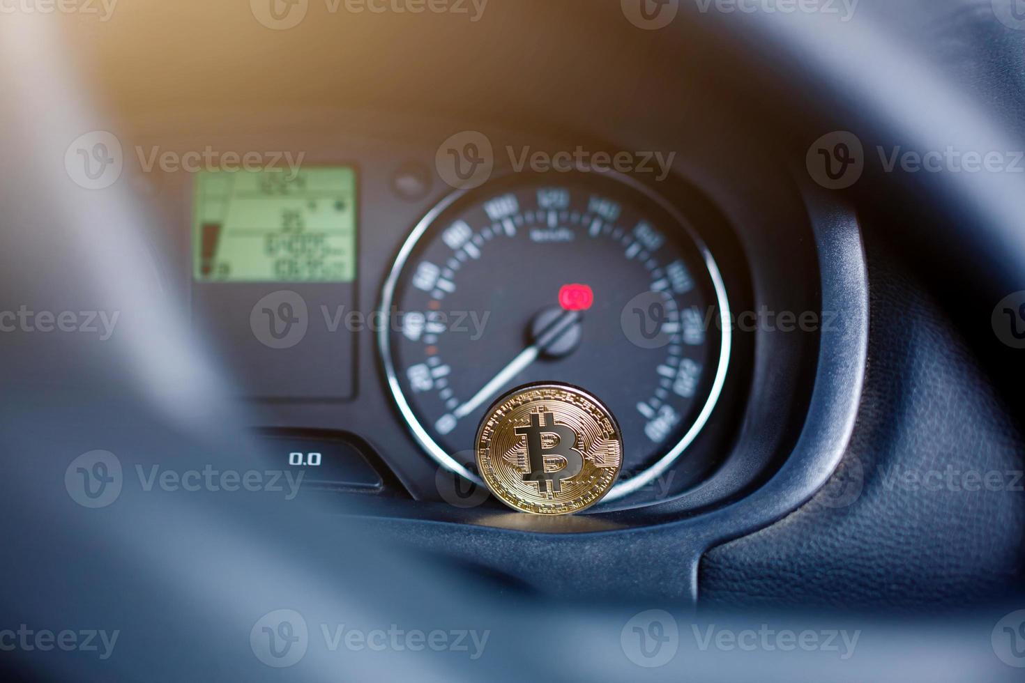 The physical coin is a gold bitcoin on the dashboard of the car next to the fuel consumption photo