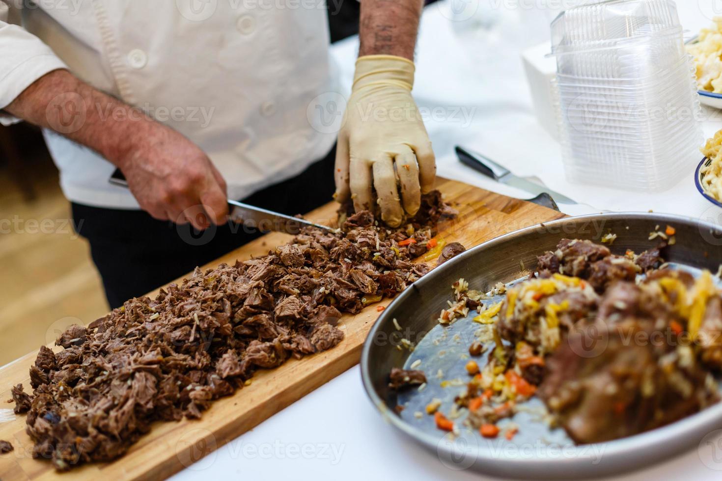 chef en cocina de hotel o restaurante vestido con delantal azul cocinando, solo manos, está cortando carne o bistec con un cuchillo grande y afilado foto