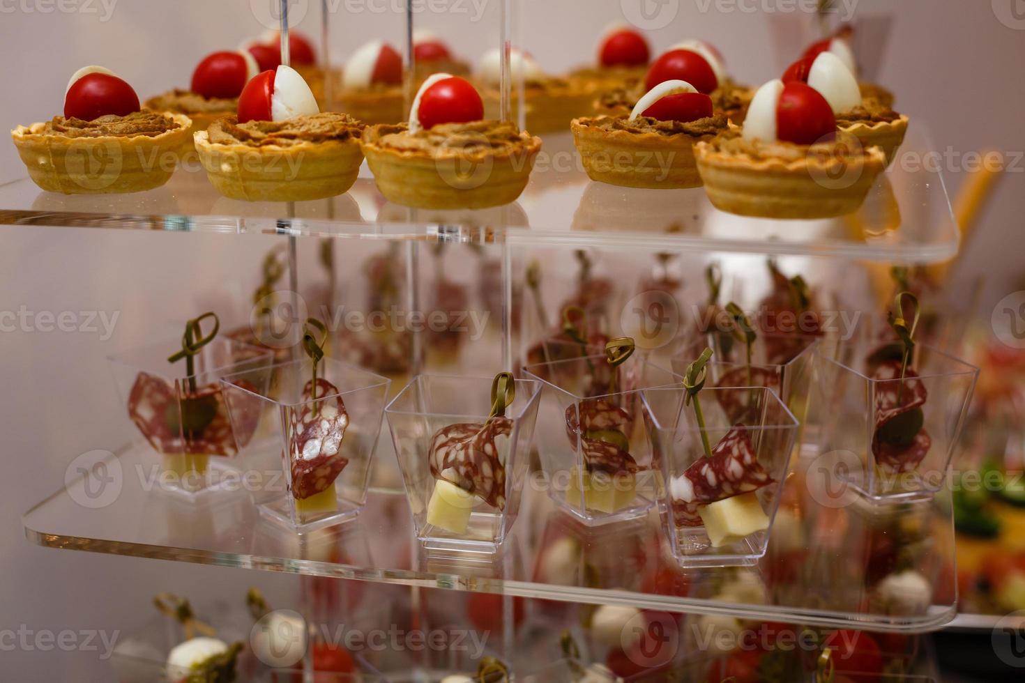 catering para fiestas. primer plano de aperitivos con tomates cherry, aceitunas verdes, aceite de oliva, queso y especias en vasos cortos sobre una mesa de madera marrón. foto