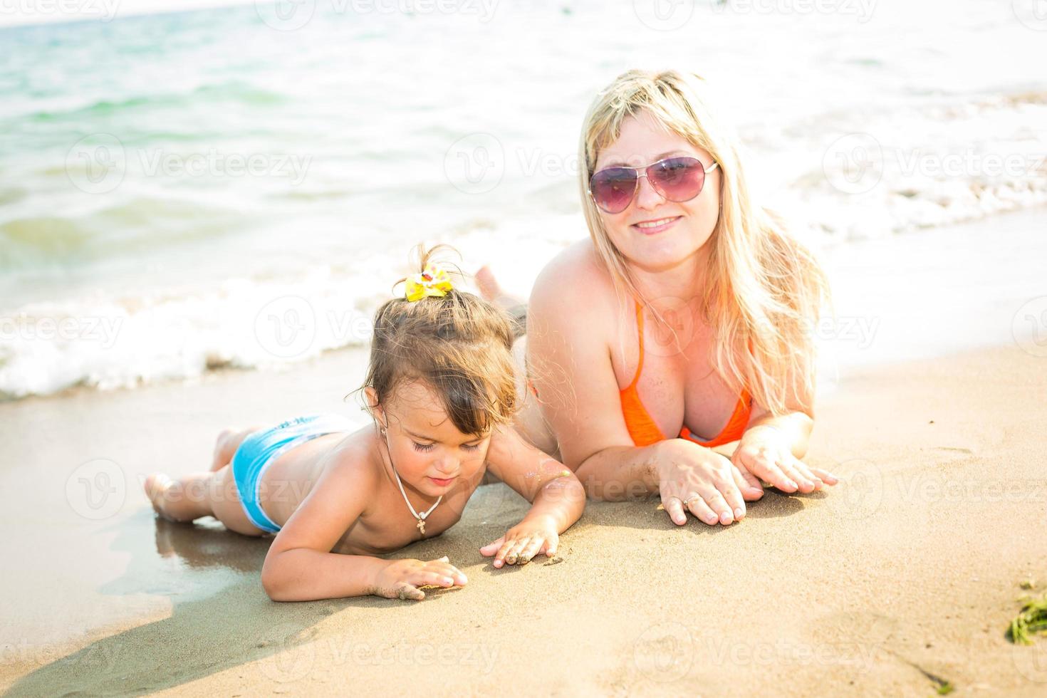 Portrait of beautiful young parents and cute little girl in swimsuits and sun glasses smiling, lying on the sunny beach. Parents are looking at their daughter photo