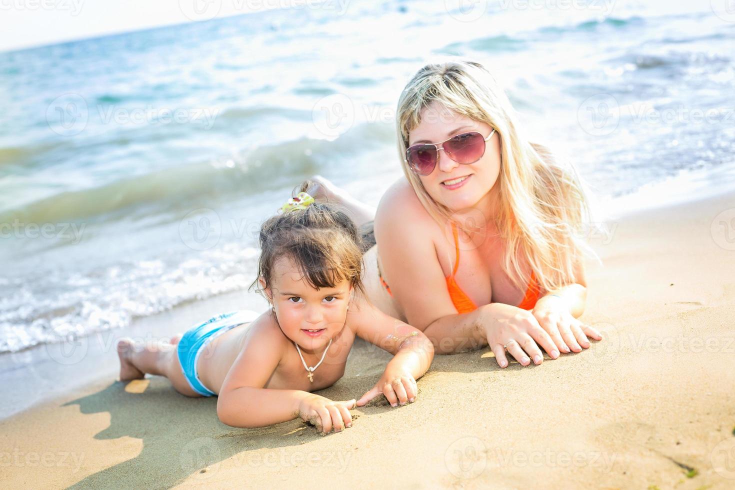 retrato de hermosos padres jóvenes y linda niñita en trajes de baño y gafas de sol sonriendo, tumbados en la soleada playa. los padres miran a su hija foto