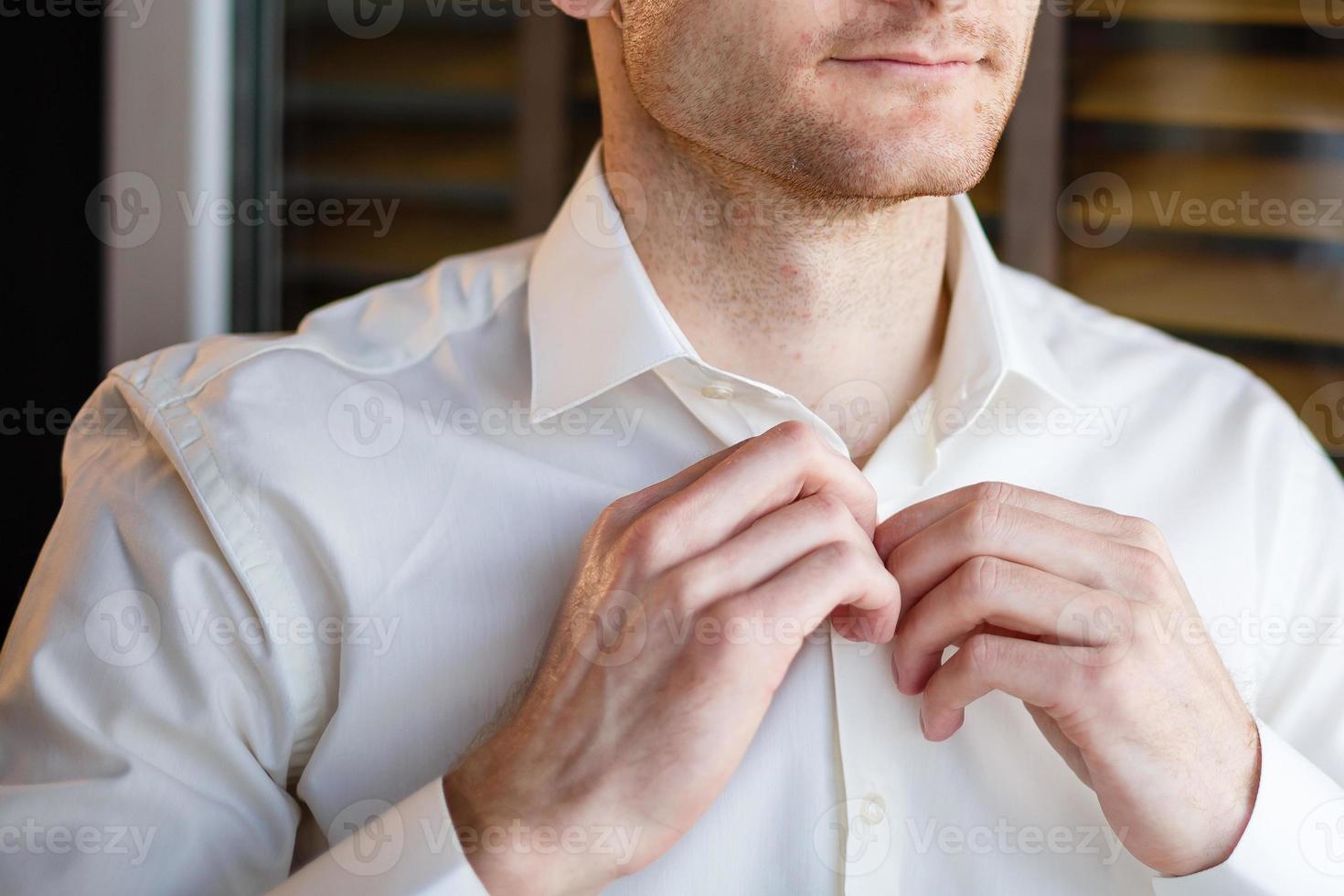 el hombre se abotona la camisa blanca parado frente a una ventana luminosa foto