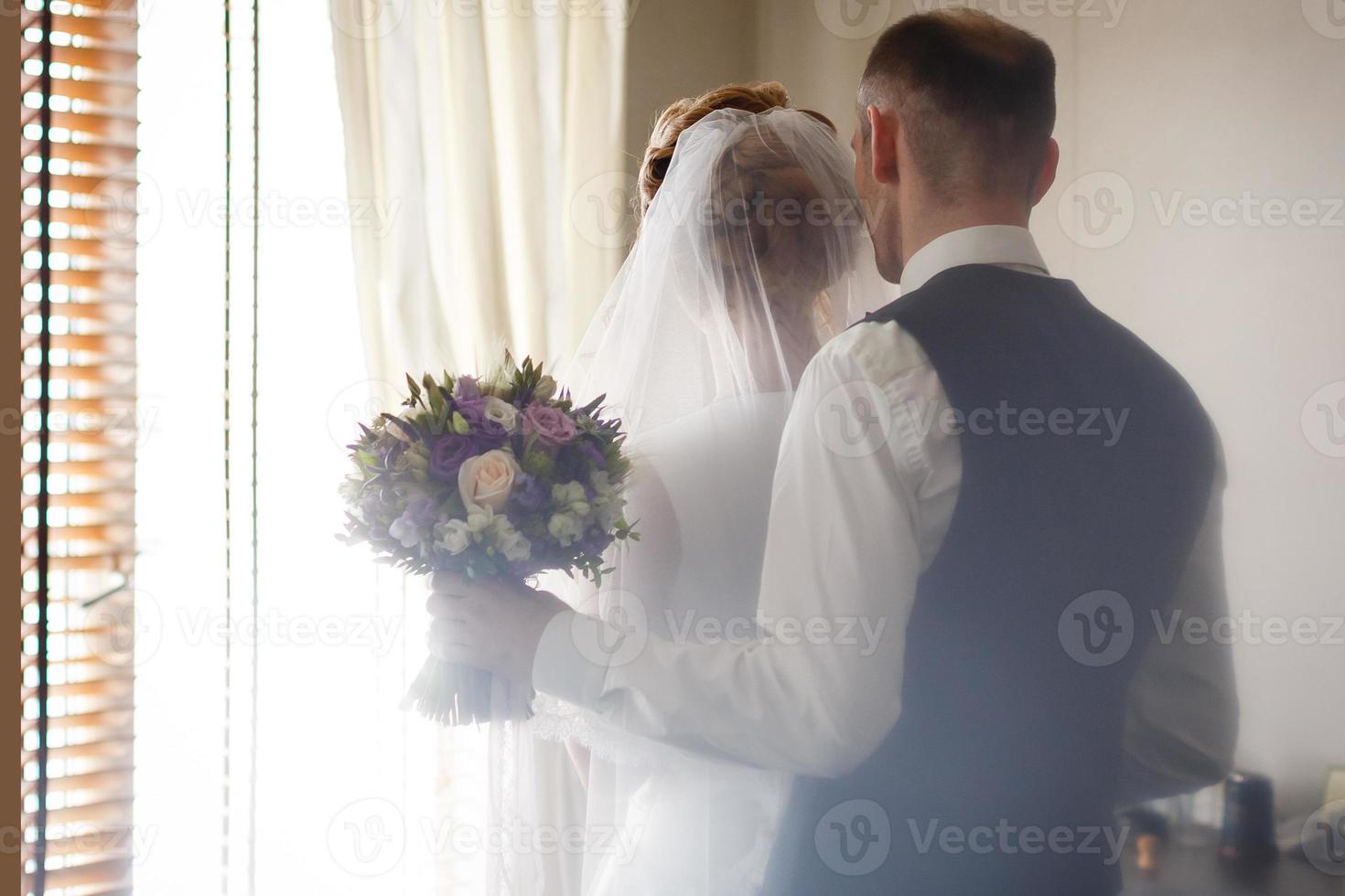 elegante novia de lujo y apuesto y elegante novio tomándose de la mano en una hermosa ventana en el fondo de un rico interior foto