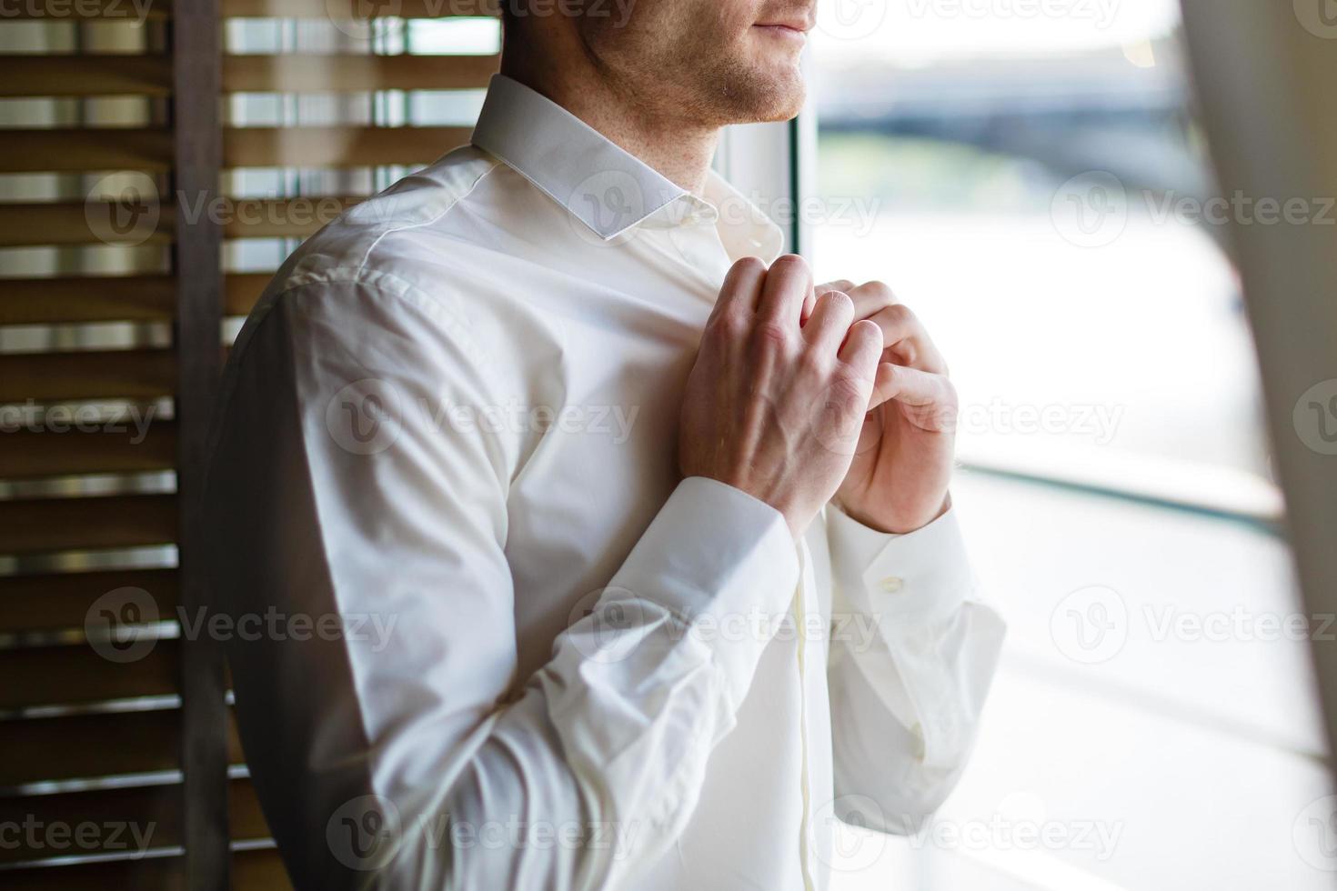el hombre se abotona la camisa blanca parado frente a una ventana luminosa foto