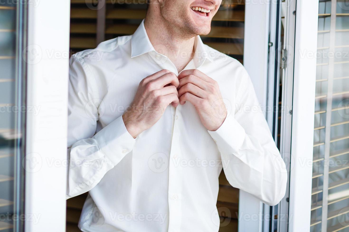 Man buttons up his white shirt standing in the front of a bright window photo