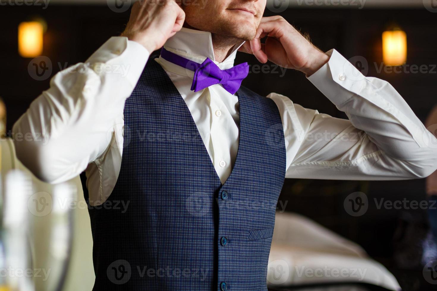 butterfly of violet color on a neck at the man. Man wears purple, purple bow-tie. The bridegroom going to the wedding. Wedding morning. The man straightens his tie. photo