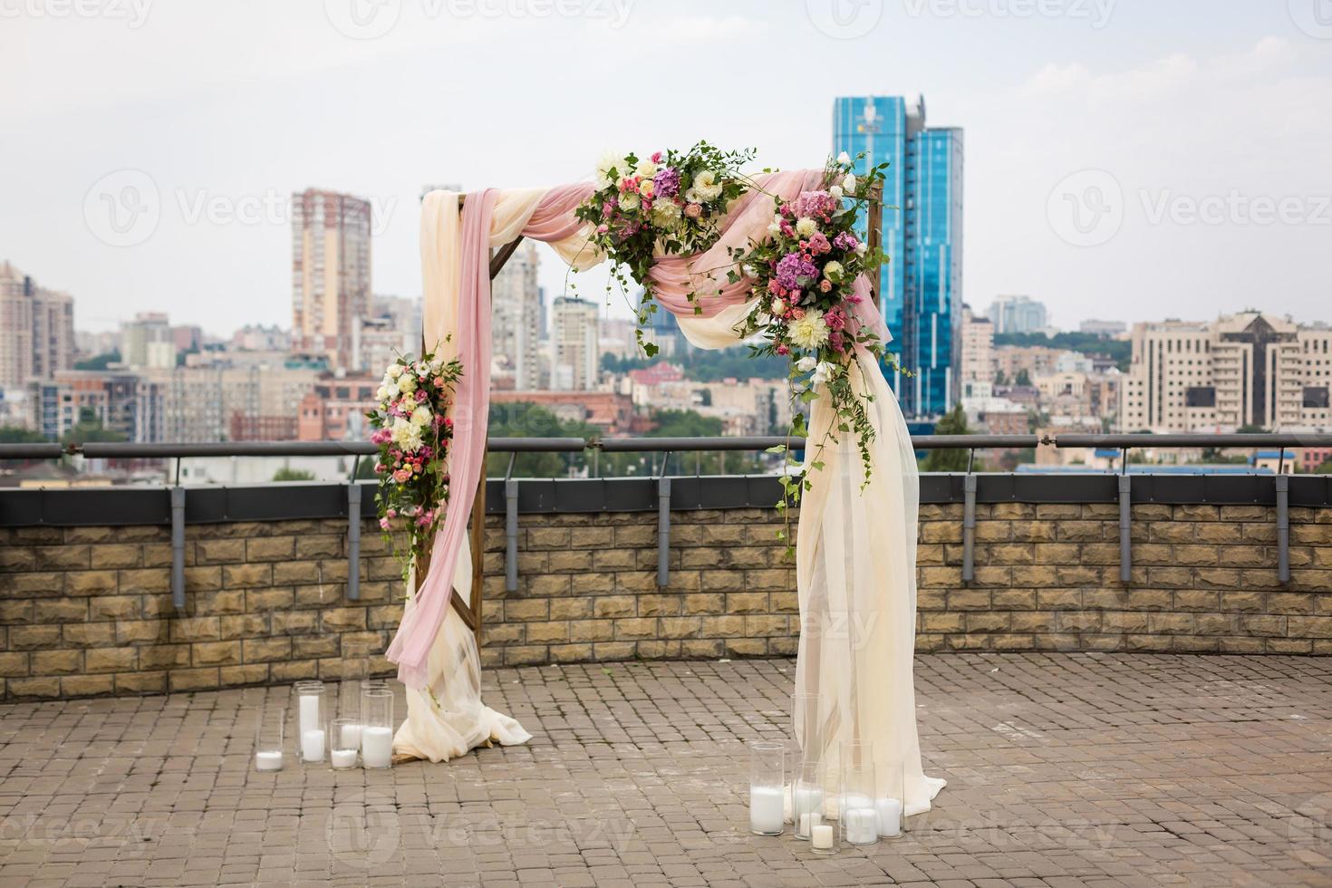 hermosa ceremonia de boda al aire libre. arco de boda hecho de tela y flores blancas y rosas en el techo con el telón de fondo de una gran ciudad. puertas antiguas, estilo rústico. foto