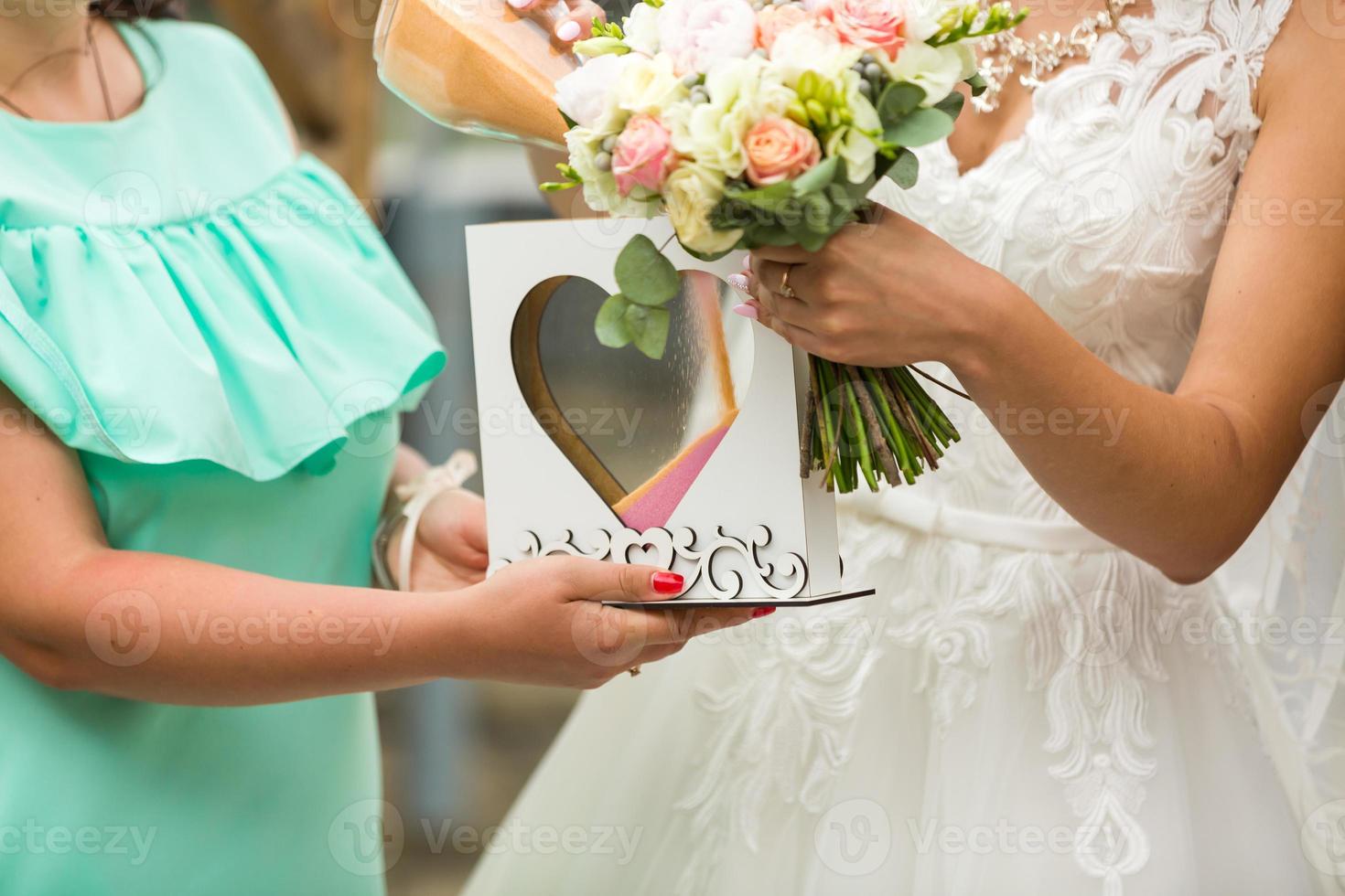 Groom and bride pouring colourful sand into box with glass heart outdoor photo