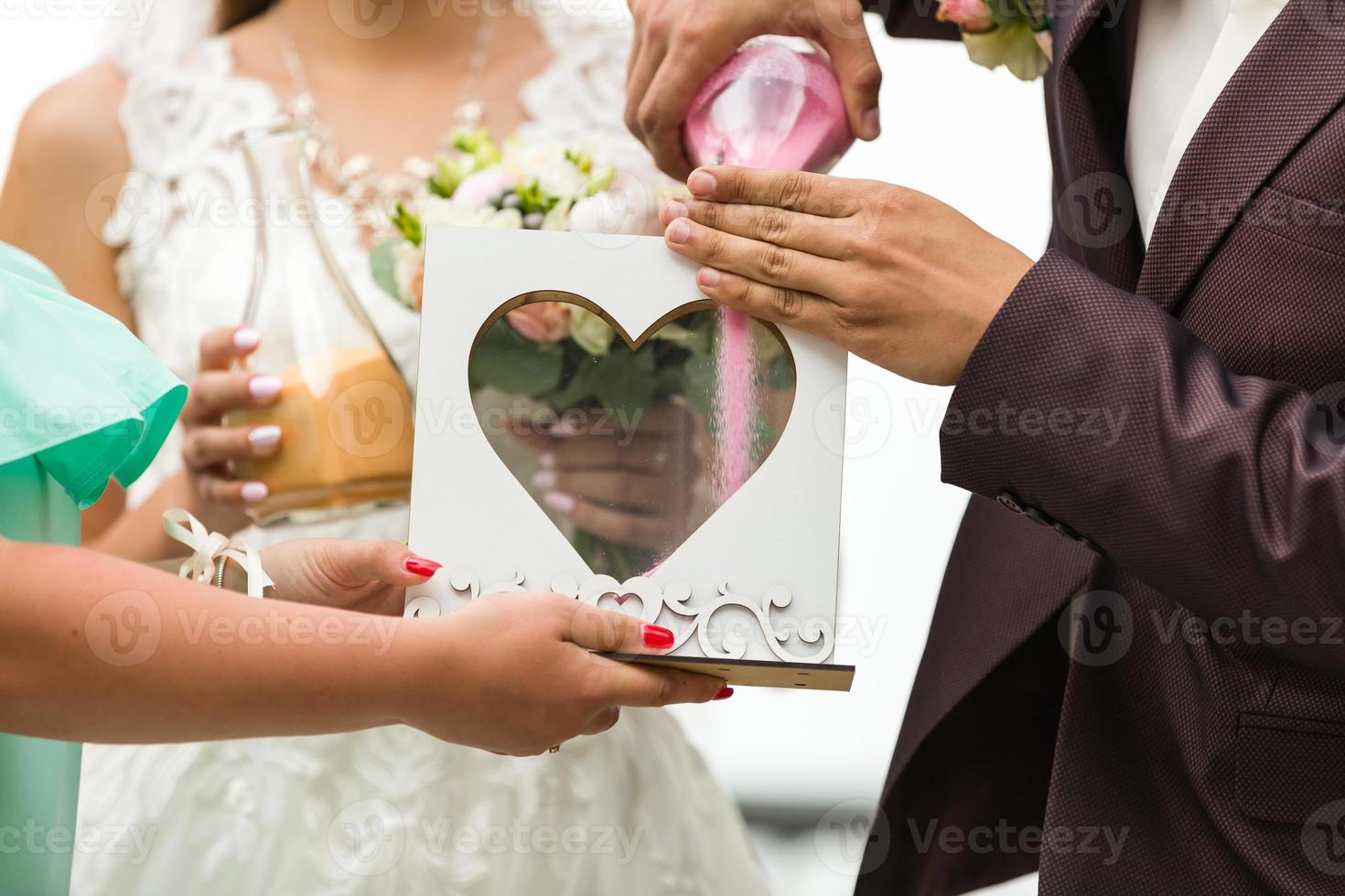 Groom and bride pouring colourful sand into box with glass heart outdoor photo