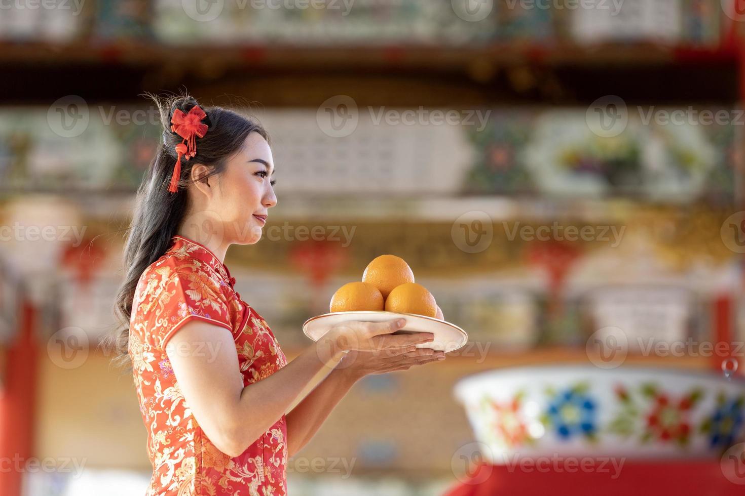 Asian woman in red cheongsam qipao dress is offering tangerine to the ancestral god inside Chinese Buddhist temple during lunar new year for best wish blessing and good luck concept photo