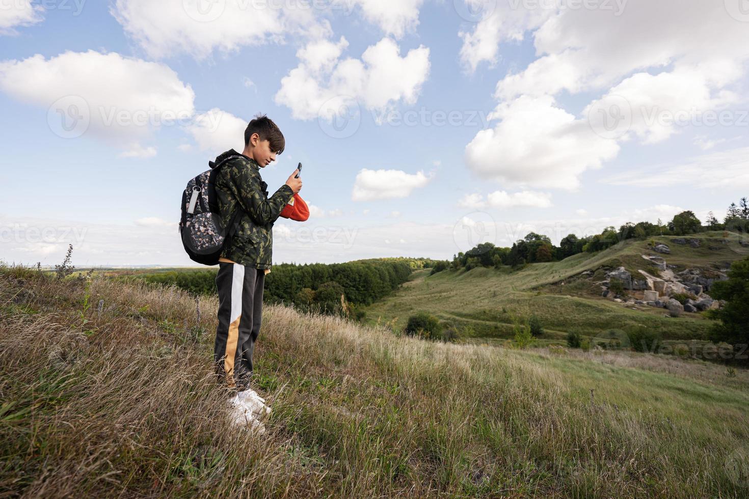 Activity on sunny autumn day, boy exploring nature. Kid wear backpack hiking and making photo on phone.