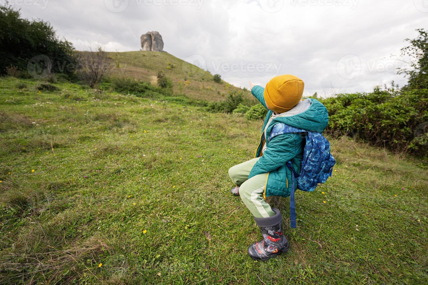 Activity on sunny autumn day, boy exploring nature. Kid wear yellow hat and backpack sit against big stone of Pidkamin, Ukraine. photo