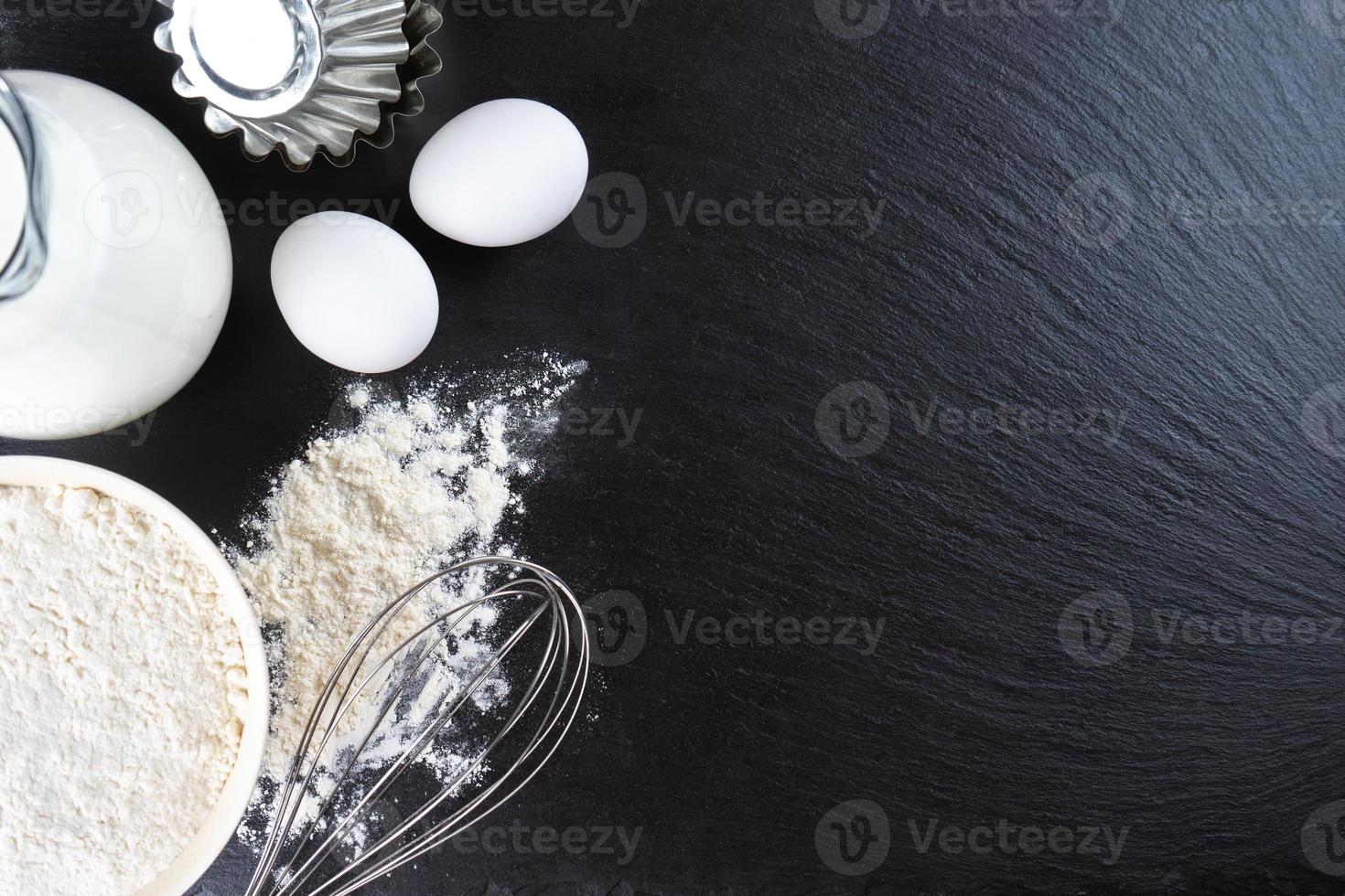 Baking ingredients flour, eggs, milk, bakeware on black slate background with copy space photo