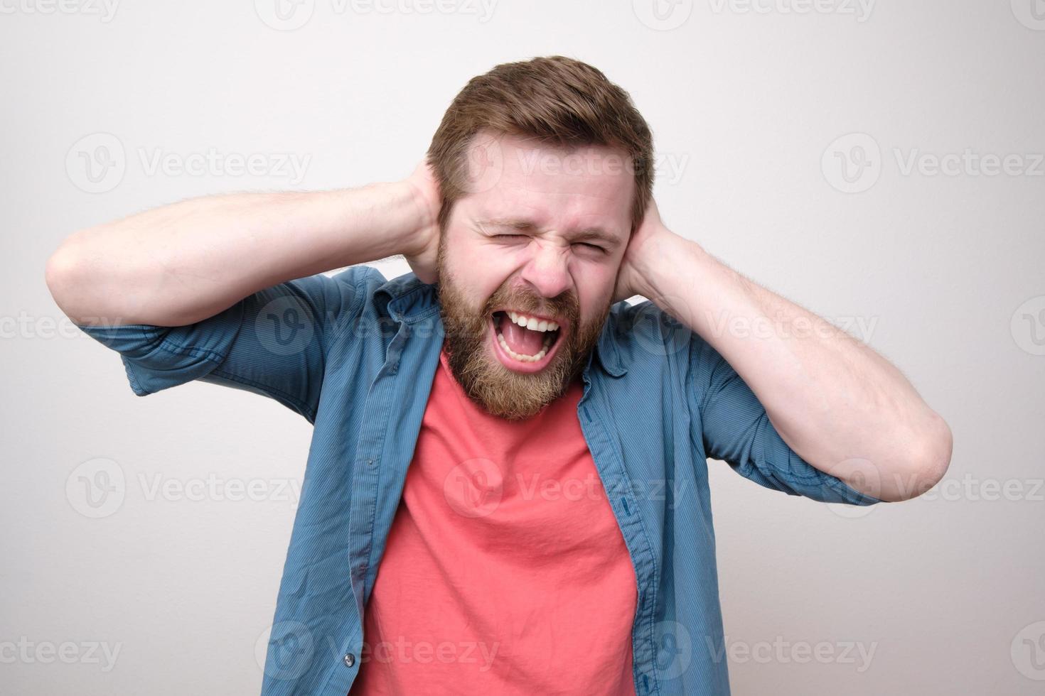 hombre caucásico estresado, está cansado del ruido, cubre los oídos con las manos y grita en voz alta. concepto de salud psicológica. foto