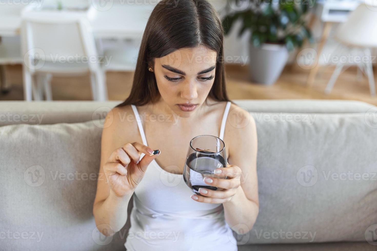 la joven toma una pastilla con un vaso de agua en la mano. mujer estresada mirando por la ventana y bebiendo medicamentos antidepresivos sedados. la mujer se siente deprimida, tomando drogas. medicamentos en el trabajo foto