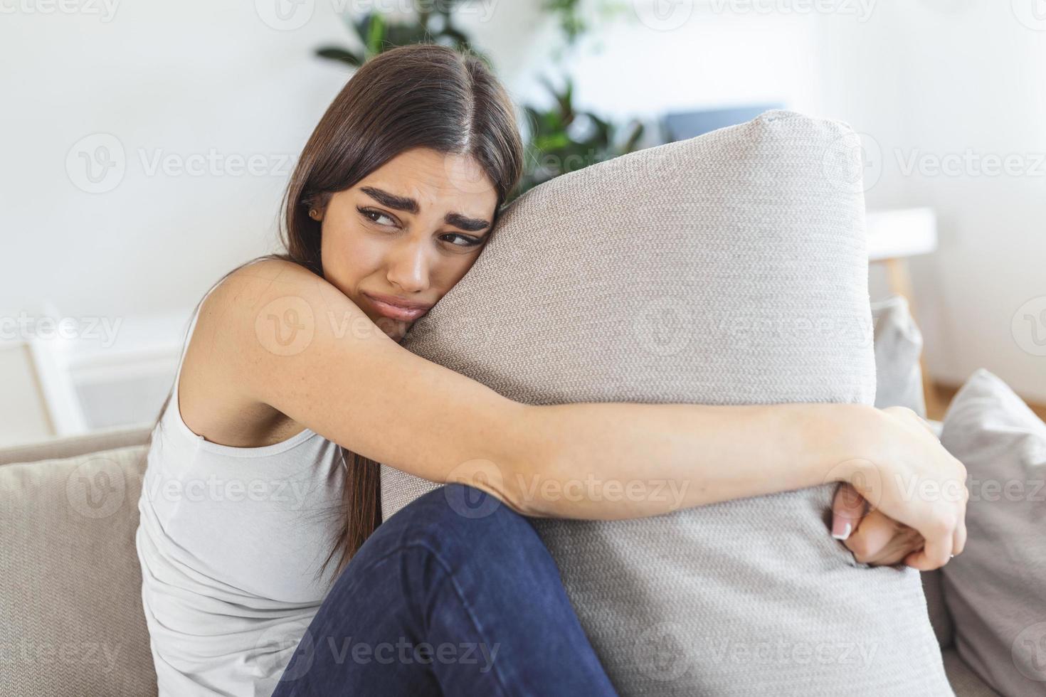 mujer joven abrazando la almohada con depresión y llorando en el sofá en casa. dama milenaria solitaria que se siente estresada y desesperanzada, sufre de trastornos del estado de ánimo o tiene problemas psicológicos foto