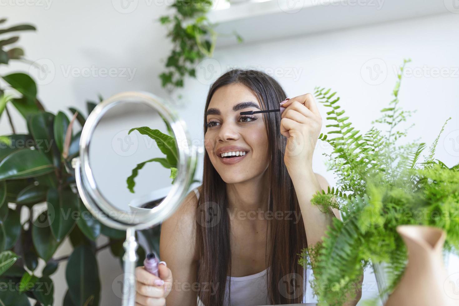 Beautiful woman using mascara on eyelash in bathroom in the morning time. Smiling young woman applying eye make up and looking at mirror. Beauty girl applying black mascara in bathroom at home. photo