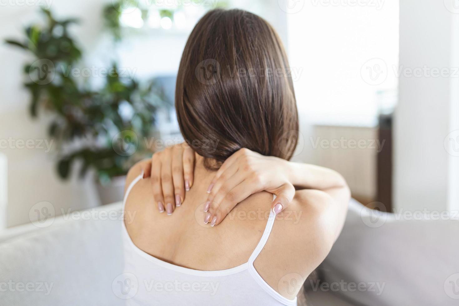 mujer joven que sufre de dolor de espalda en casa. retrato de una joven morena sentada en el sofá de casa con dolor de cuello y dolor de espalda. bella mujer con dolor de columna o de cuello foto