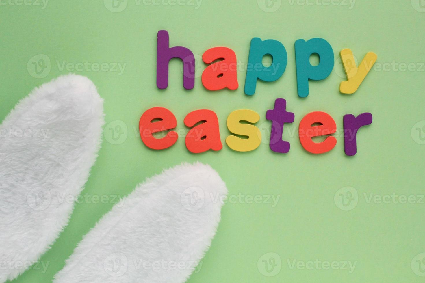 Happy Easter colored lettering and white bunny fur ears on green background. Holiday spring composition creative concept. Idea for funny event postcard, banner, poster, party. Top view, flatlay photo
