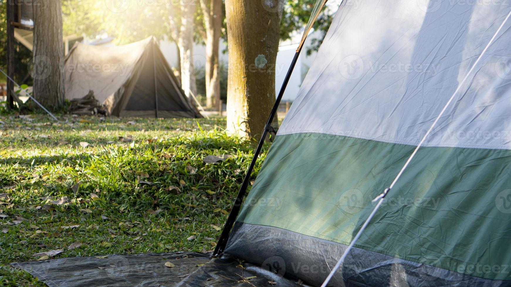 A corner of a green tent spread out on a black canvas under the shade of a big tree. Background were lawns and other tents in the distance under the morning light. photo