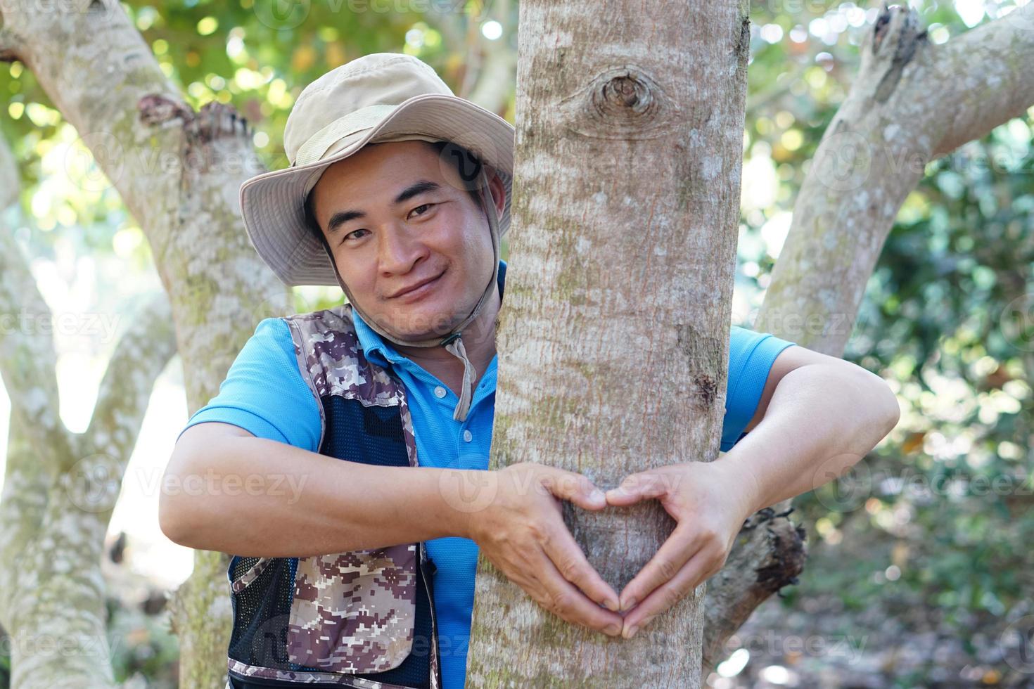 un apuesto botánico asiático abraza el tronco de un árbol en el bosque. hacer manos en forma de corazón. concepto, amor a la naturaleza y protección, bosque, ecología y conservación del medio ambiente. foto