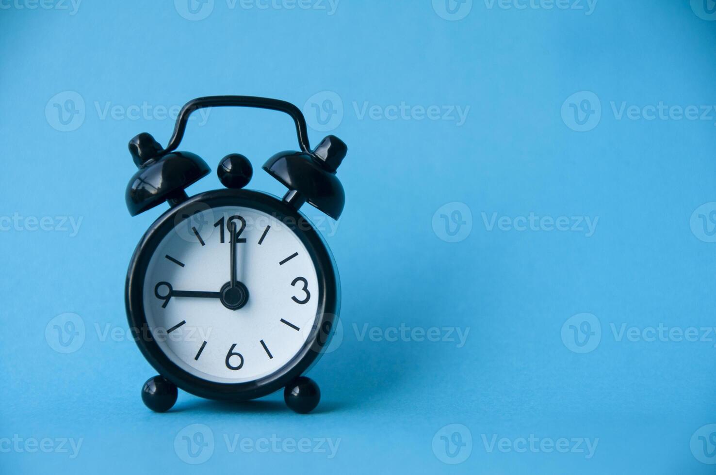 Black alarm clock isolated on light blue background. The clock set at 9 o'clock. photo