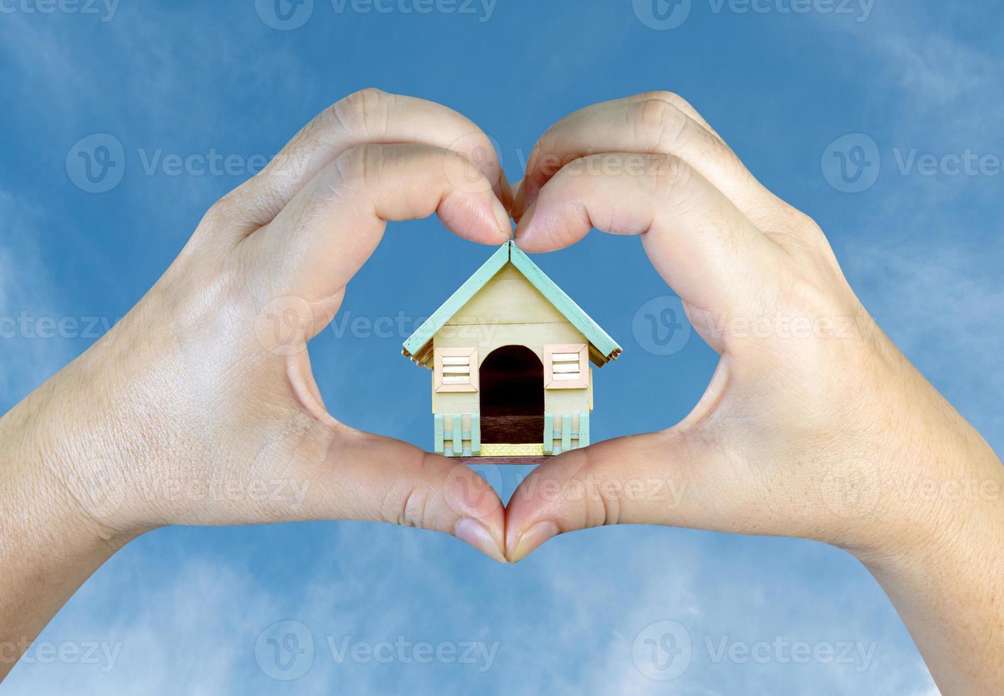 person hands making a heart shape with wooden house on sky background photo