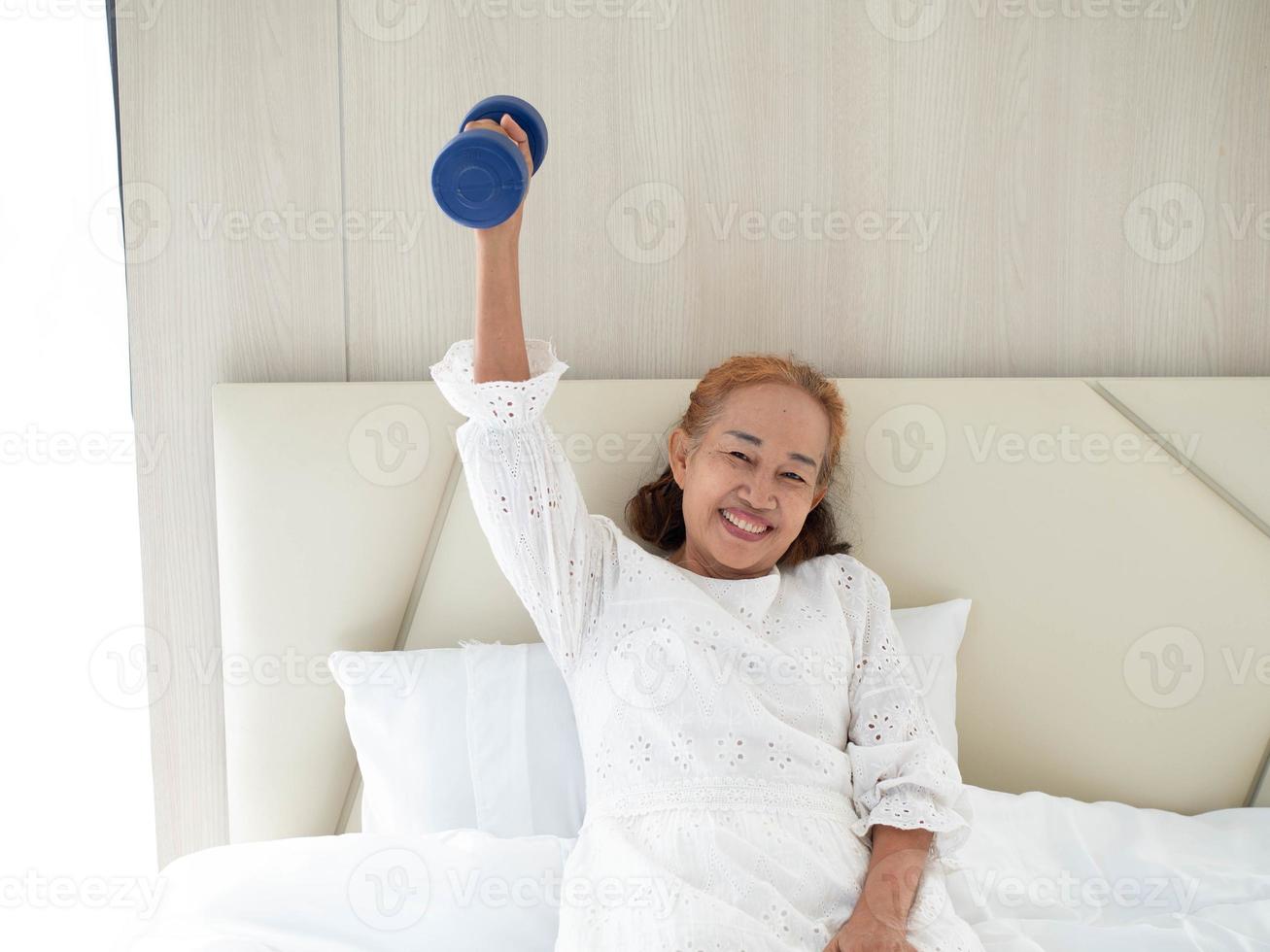 Female senior older woman lady her person portrait look at camera holding dumbbell happy smile indoor bedroom home house strong healthy happy lifestyle activity exercise workout vitality wellness photo