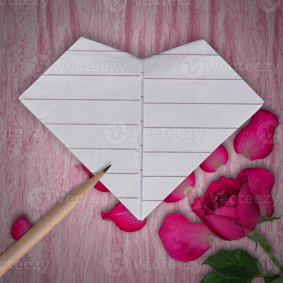 Origami paper white heart, pencil and rose pink flower on wooden table background photo