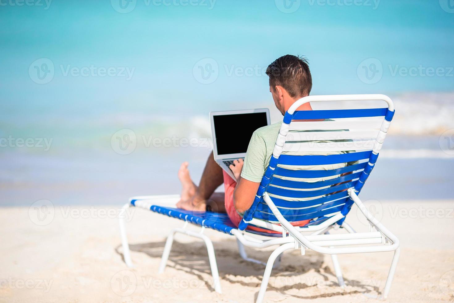 Young man with laptop on tropical beach. Man sitting on the chaise-lounge with computer photo