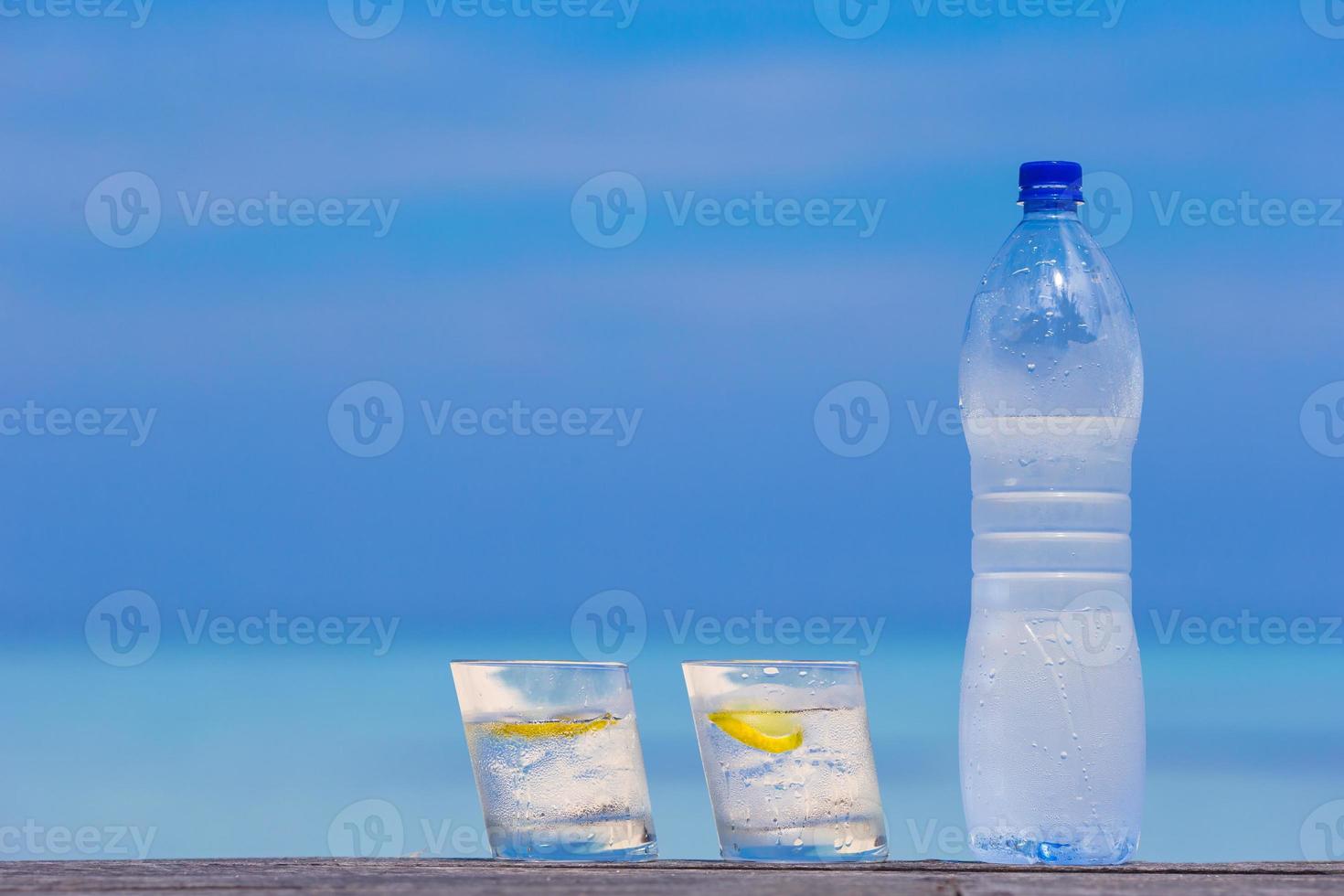 Glasses of waters with lemon and bottle on wooden on sea background photo