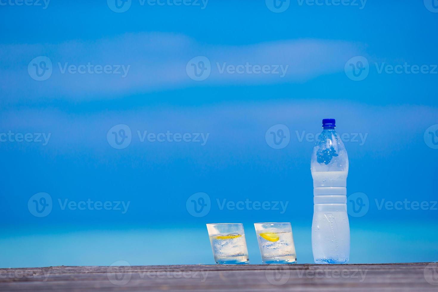Glasses of waters with lemon and bottle on wooden on sea background photo