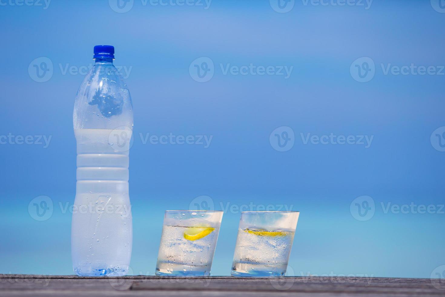 Glasses of waters with lemon and bottle on wooden on sea background photo