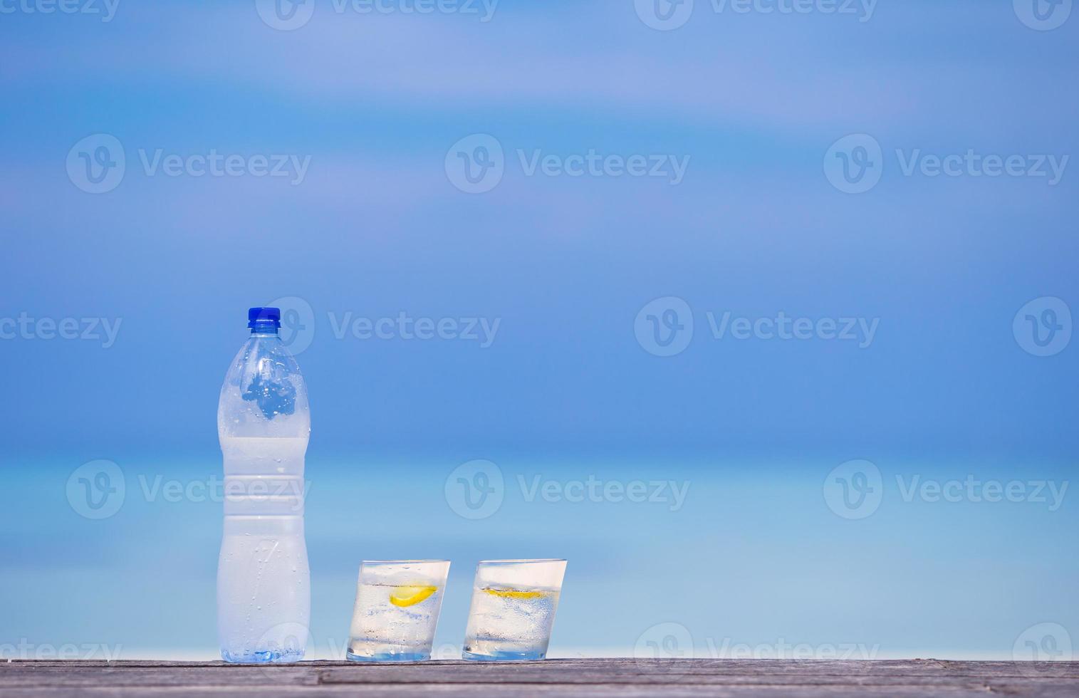 Glasses of waters with lemon and bottle on wooden on sea background photo