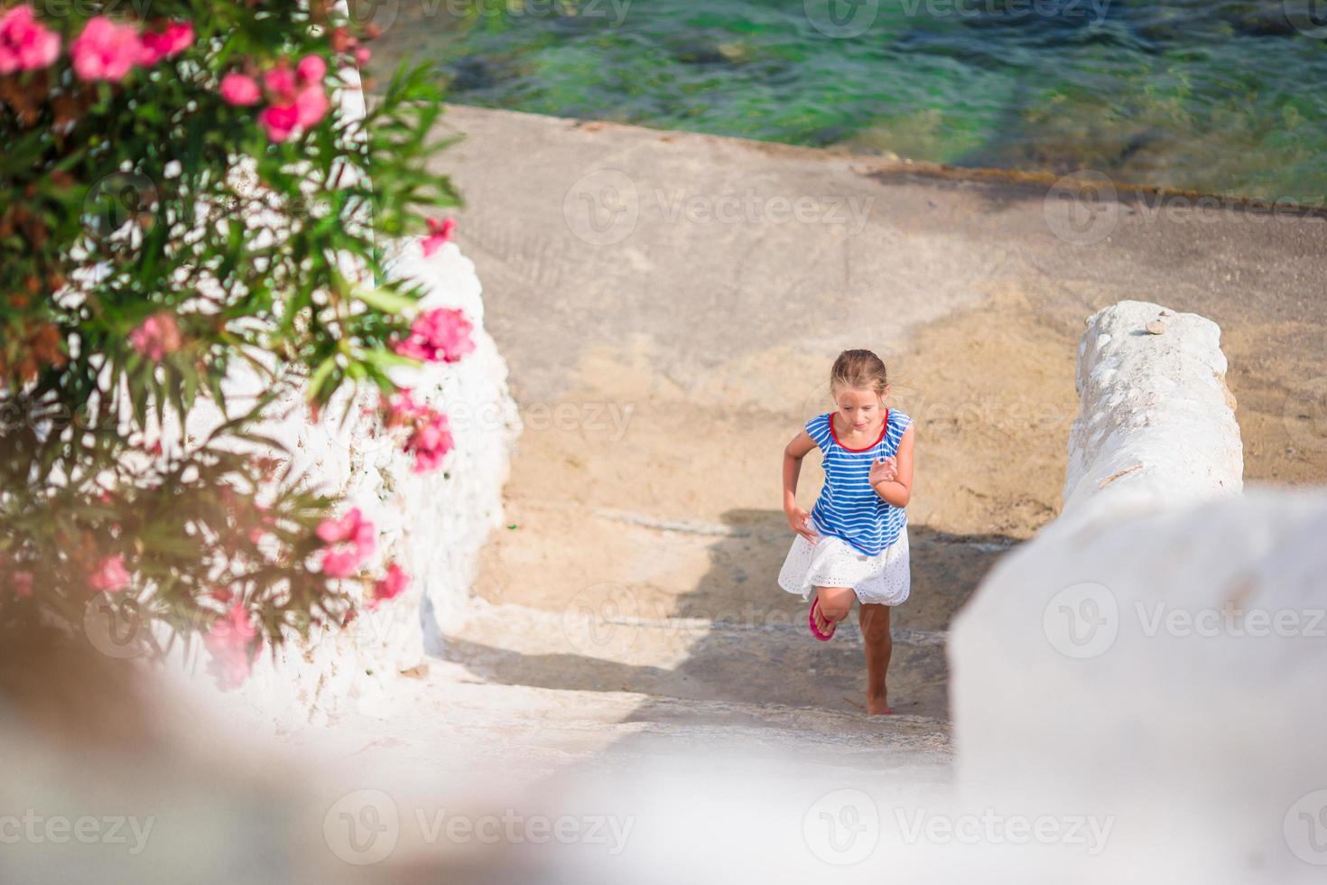 Adorable girl having fun outdoors. Kid at street of typical greek traditional village with white walls and colorful doors on Mykonos Island, in Greece photo