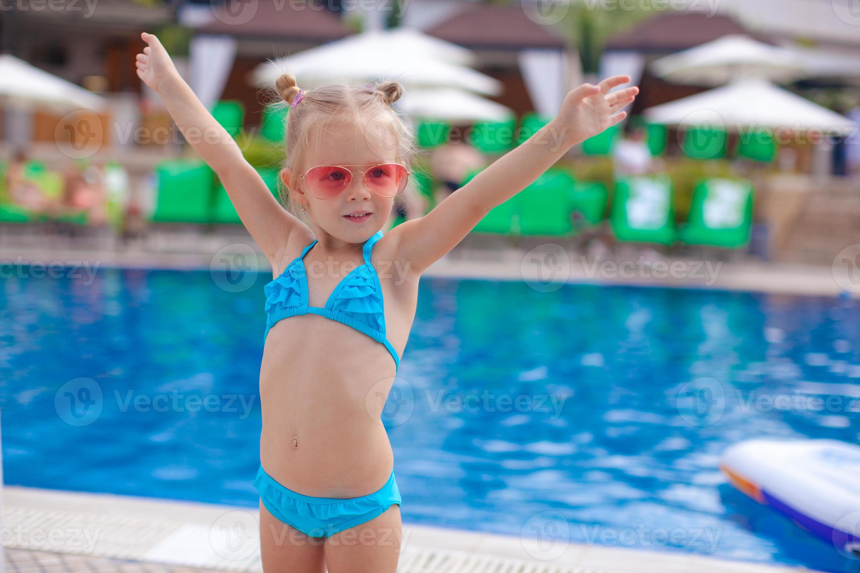 Niña linda en traje de baño azul sosteniendo una tabla de natación de  plástico rosa mientras está de pie frente a la piscina y pone los dedos de  los pies en el