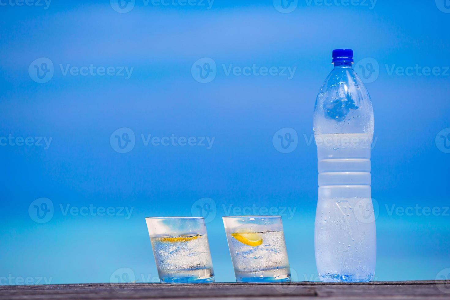 Glasses of waters with lemon and bottle on wooden on sea background photo