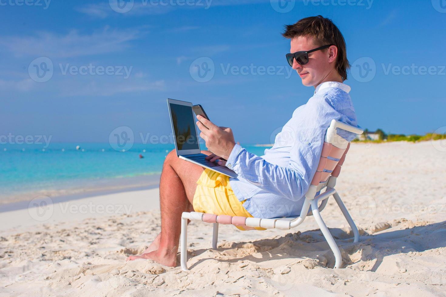 Young businessman using laptop and telephone on tropical beach photo