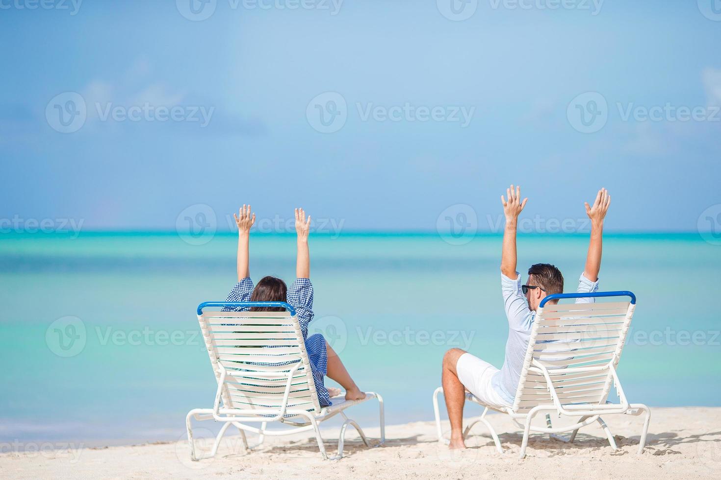 Two happy people having fun on the beach, sitting on comfortable sunbed with raised up hands for joy photo