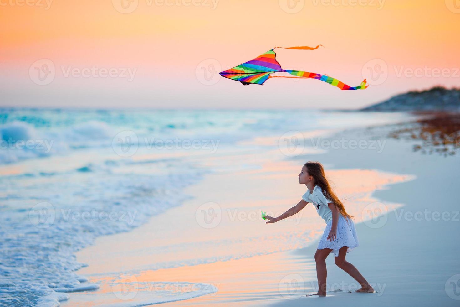 Little running girl with flying kite on tropical beach at sunset. Kids play on ocean shore. Child with beach toys. photo