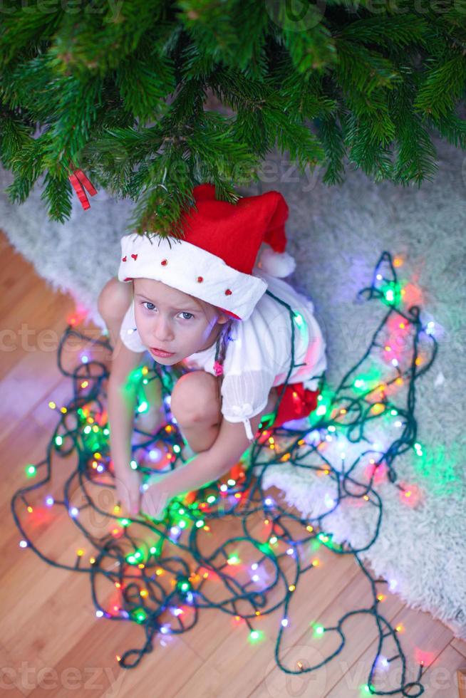 Little beautiful girl in Santa Claus hat sitting under the Christmas tree among garlands photo