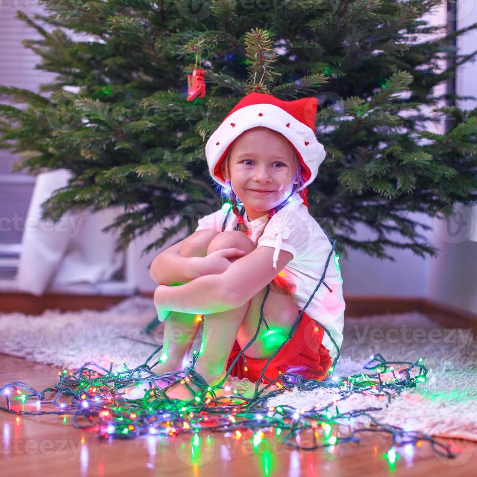 Little beautiful girl in Santa Claus hat sitting under the Christmas tree among garlands photo