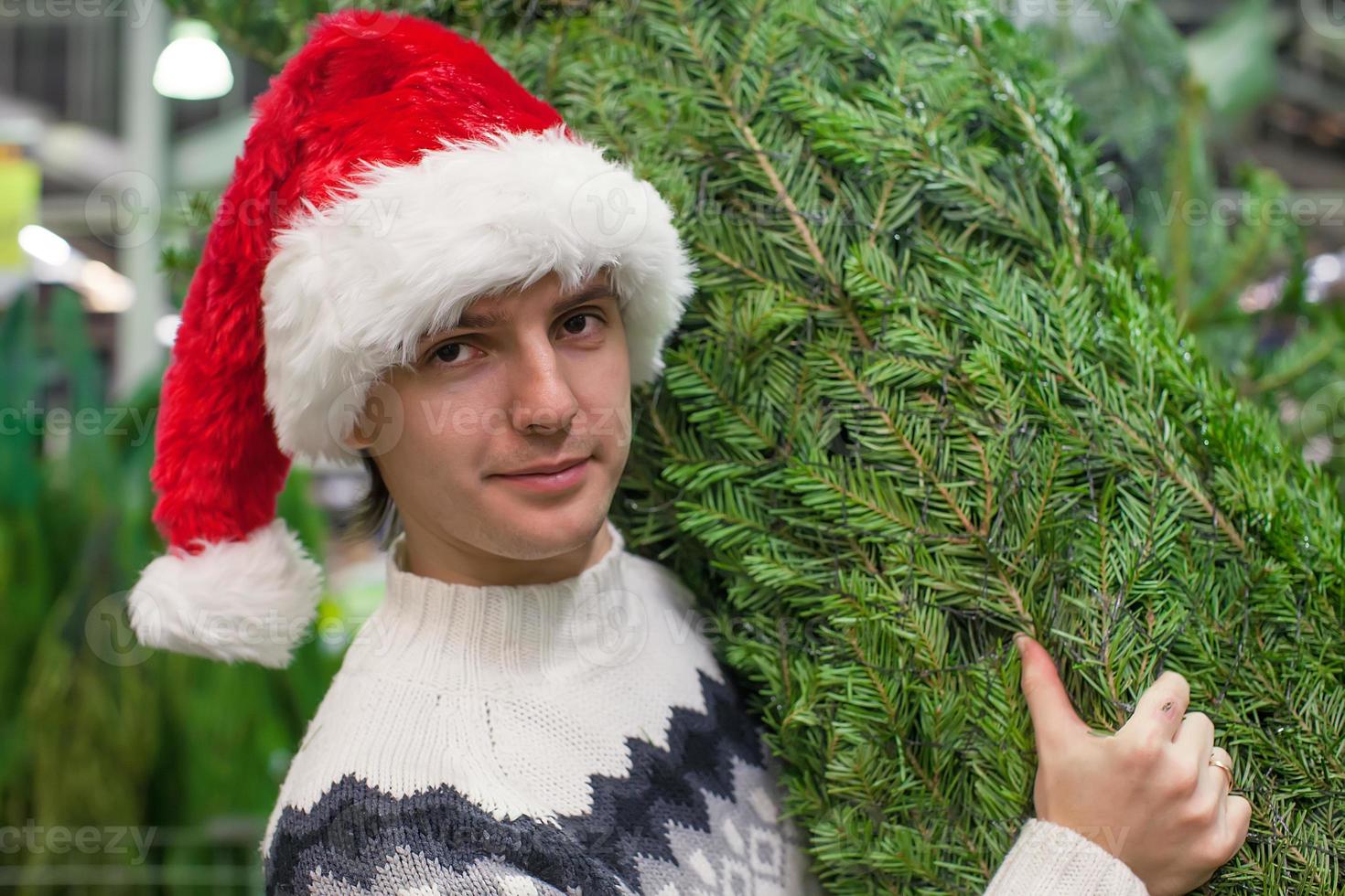 retrato de un joven con sombrero de santa comprando un árbol de navidad y mostrando los pulgares hacia arriba foto