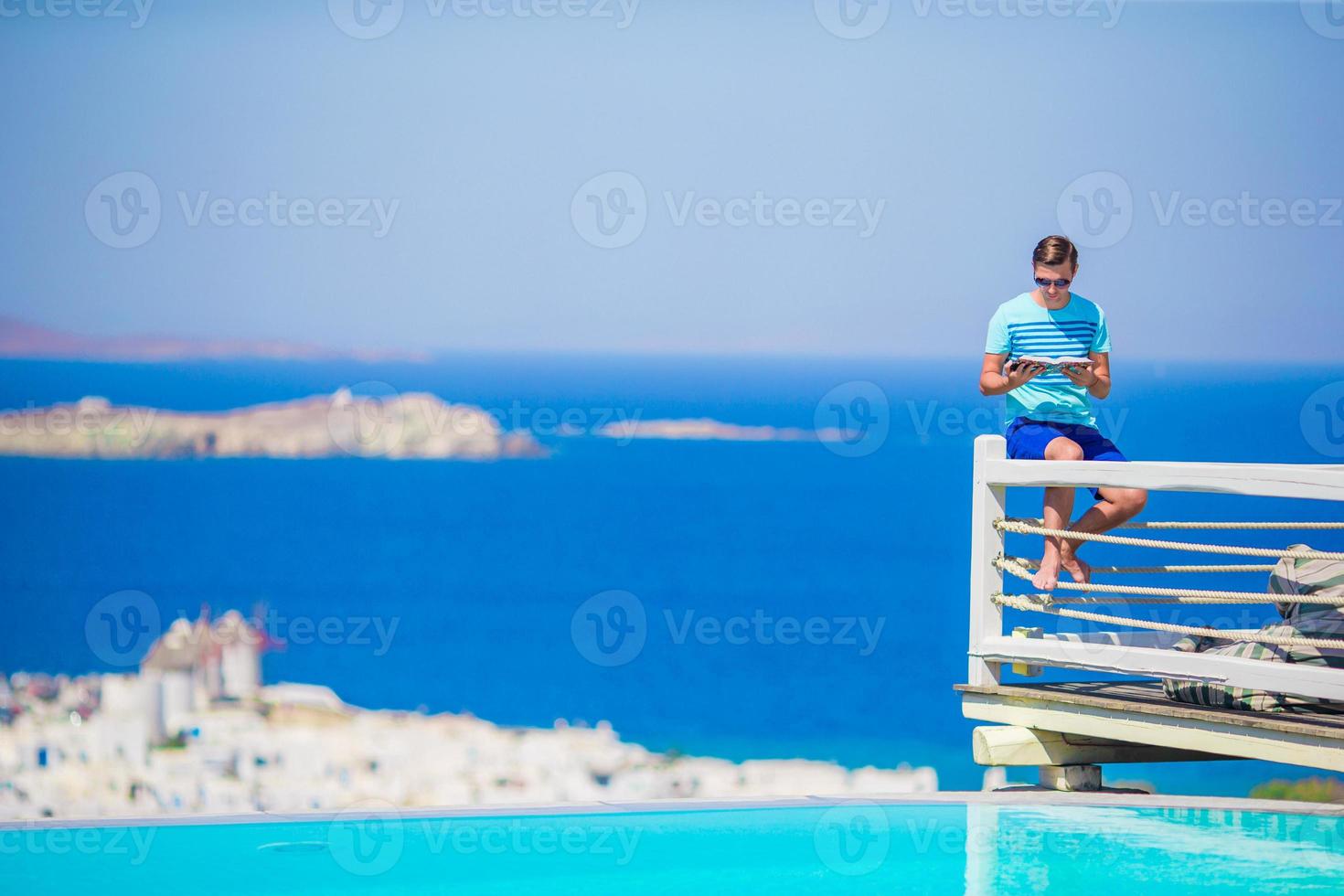 Young man relaxing near pool with amazing view and reading book in Greece. Beautiful famous background photo