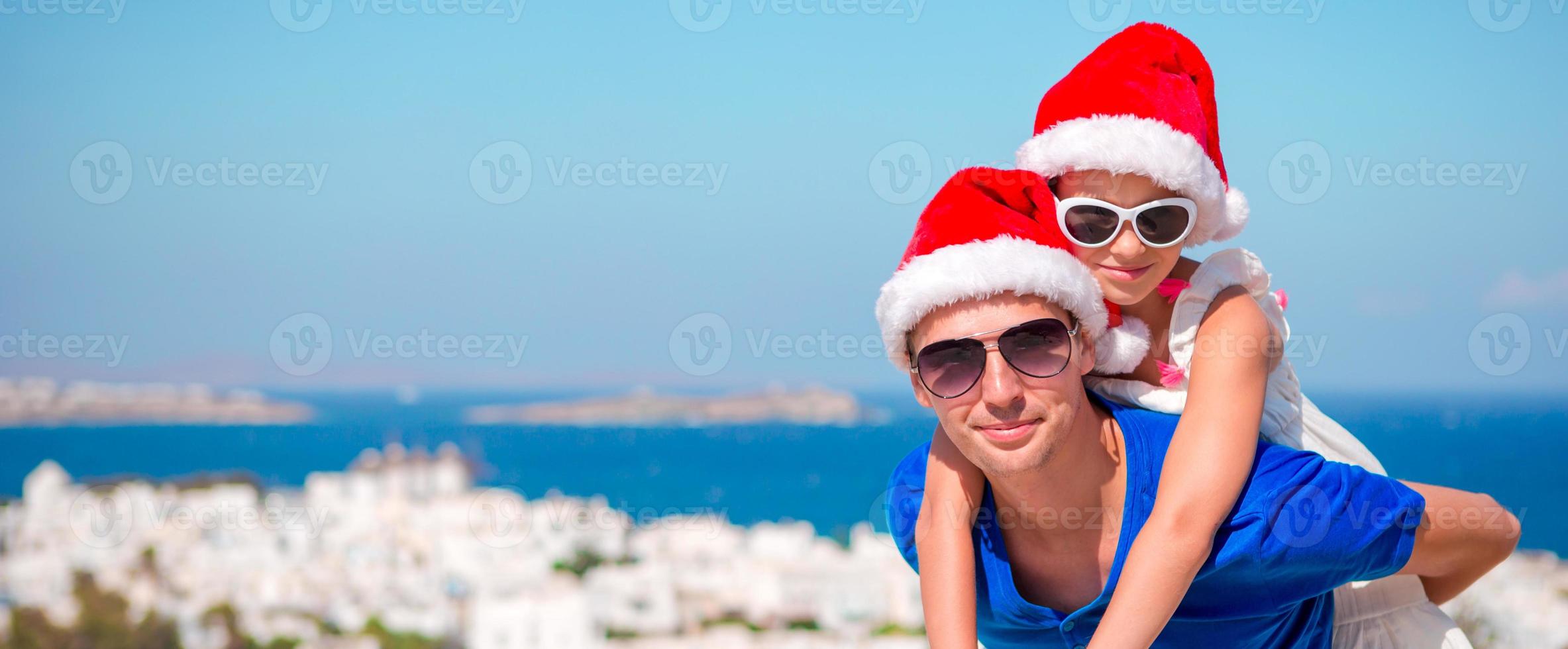 retrato de niña y papá feliz en sombreros de santa disfrutan de vacaciones de navidad con hermosa vista. panorama en el antiguo pueblo de mykonos en grecia. foto